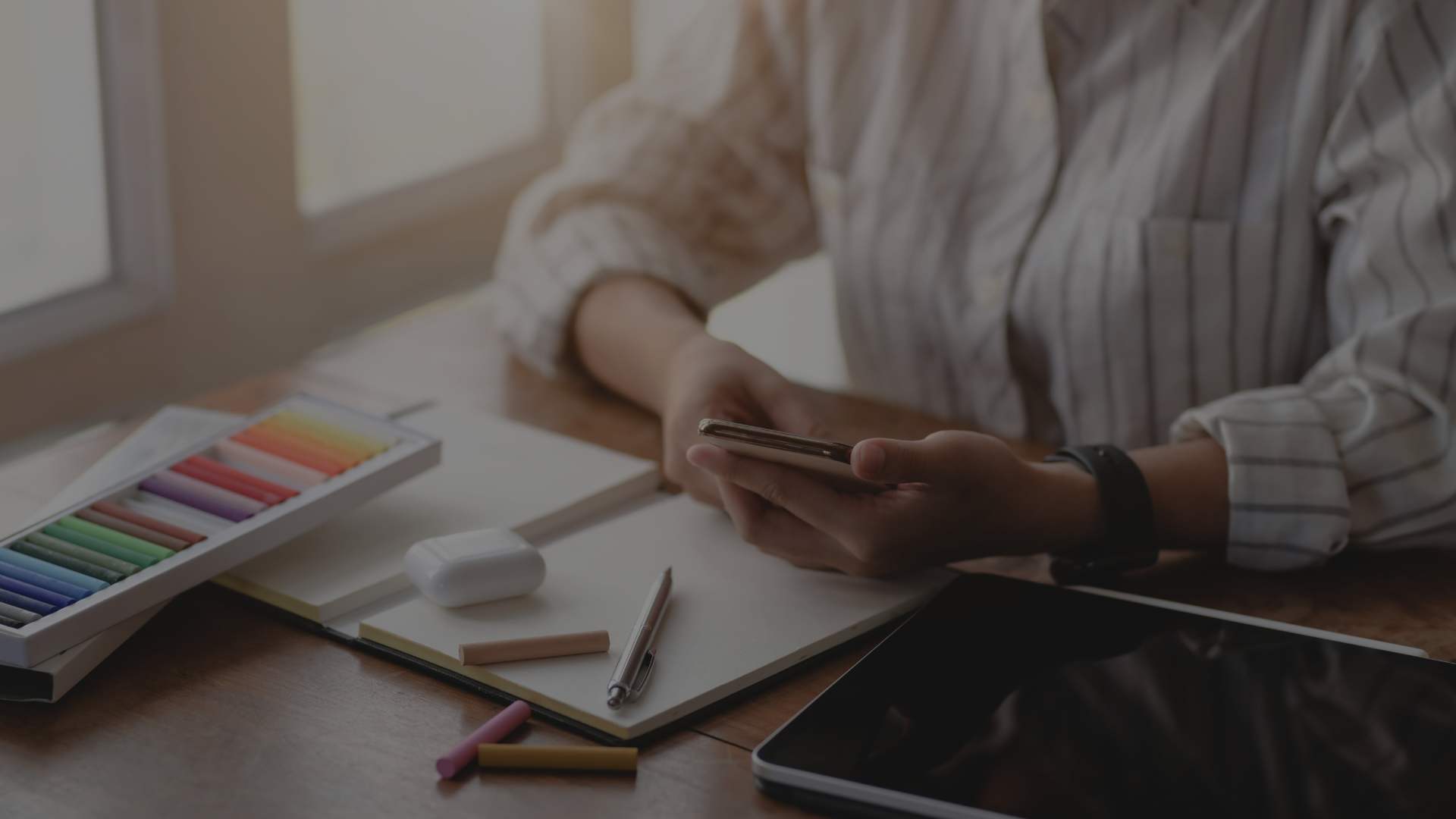 Junge Frau sitzt an einem Schreibtisch, hält ein Smartphone, vor ihr liegen Buntstifte und Schreibutensilien – symbolisiert Kreativität