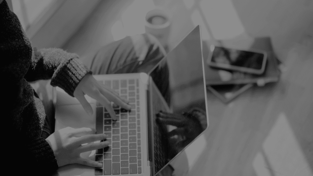 Schwarz-weiß Bild einer Frau, die auf dem Boden sitzt, ein MacBook auf dem Schoß hat und auf der Tastatur tippt – Symbolbild für Marketingkommunikation.