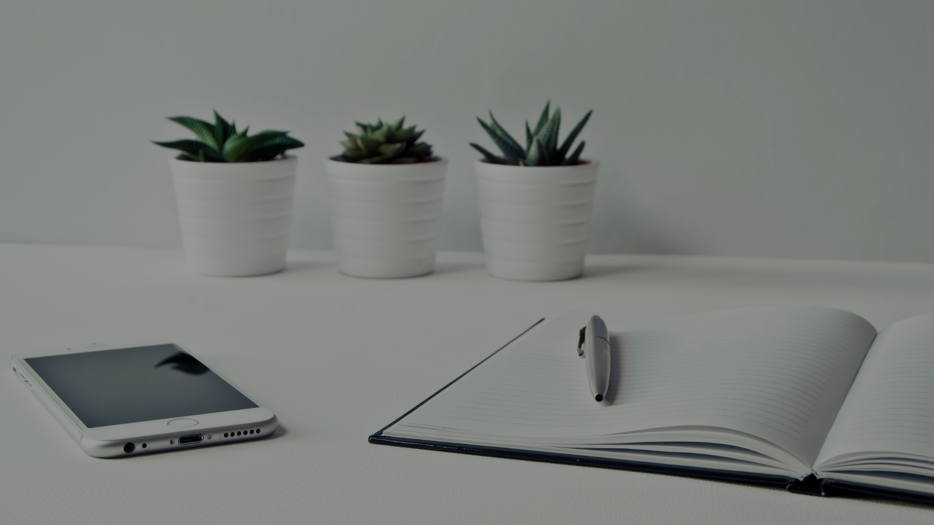 Weißer Schreibtisch mit Notizbuch, Stift und Smartphone – im Hintergrund drei kleine weiße Töpfe mit Pflanzen, symbolisiert Organisation und Struktur.
